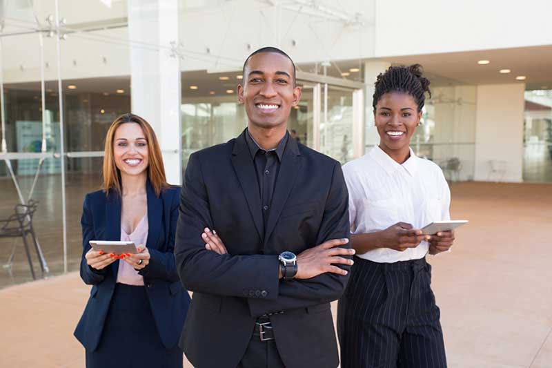 Três profissionais sorridentes em trajes sociais, em frente a um prédio corporativo de vidro. Ao centro, um homem negro de terno preto com os braços cruzados; à esquerda, uma mulher branca segurando um tablet; à direita, uma mulher negra com camisa branca e calça listrada, também segurando um tablet